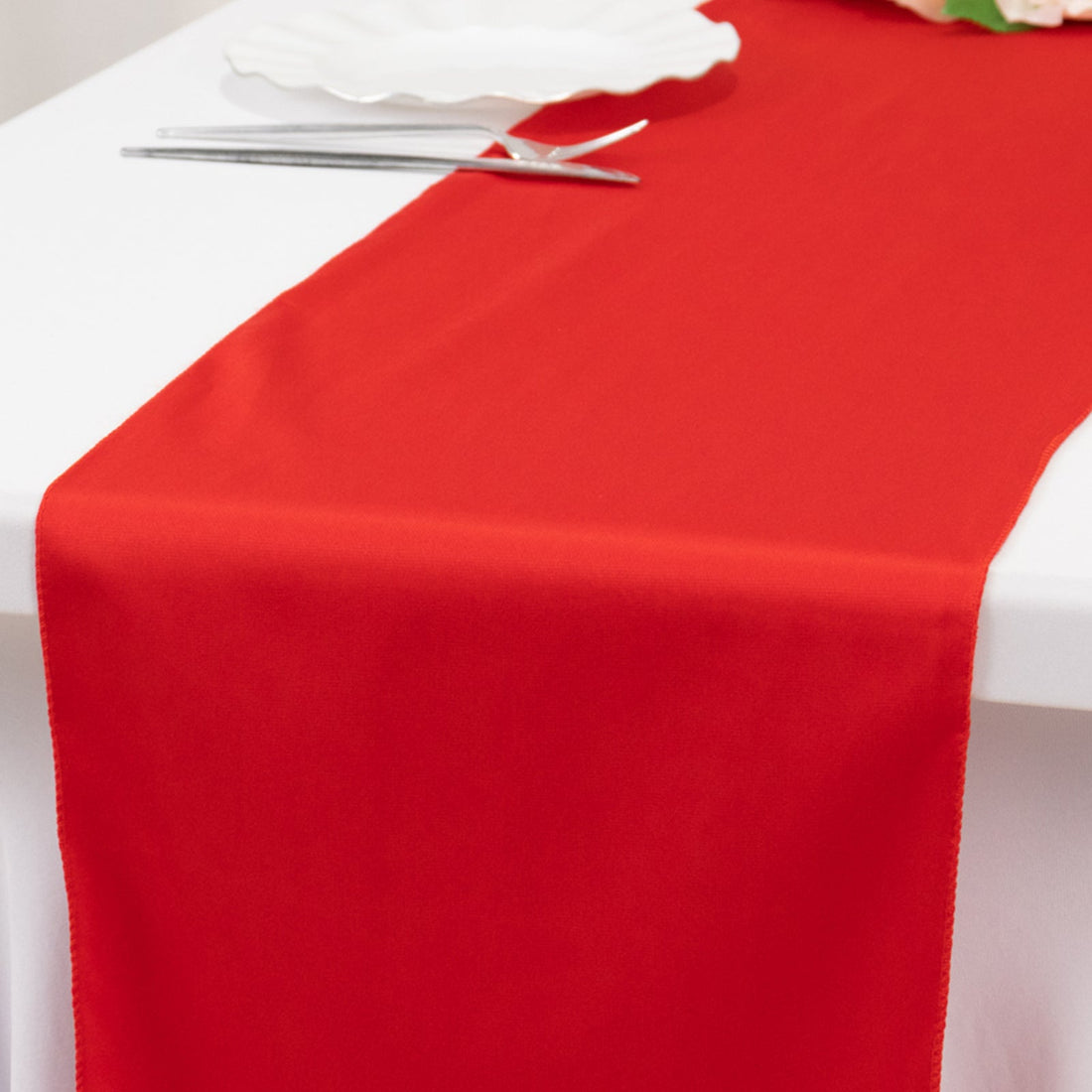 Red table runner on a white table setting with utensils and a plate.