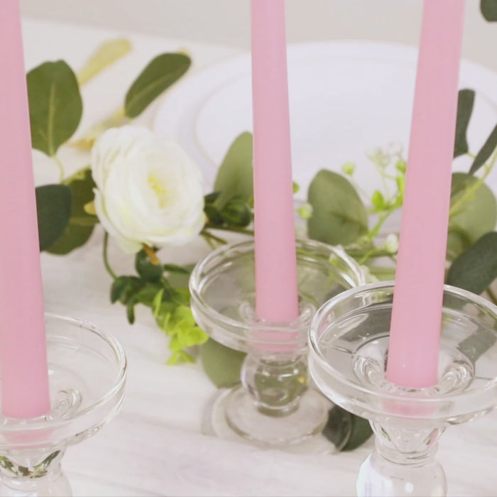 Pink taper candles in glass holders with greenery and a white flower.