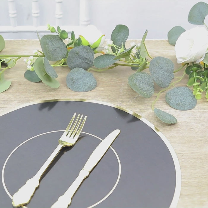 Black round placemat with gold rim on a wooden table with greenery and flowers.