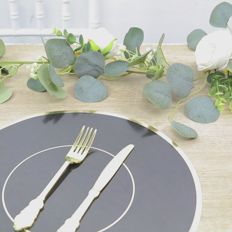Black round placemat with gold rim on a wooden table with greenery and flowers.