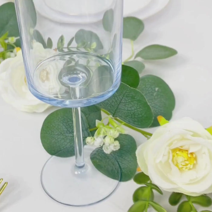 Transparent dusty blue wine glass with long stem on a table with greenery and flowers.