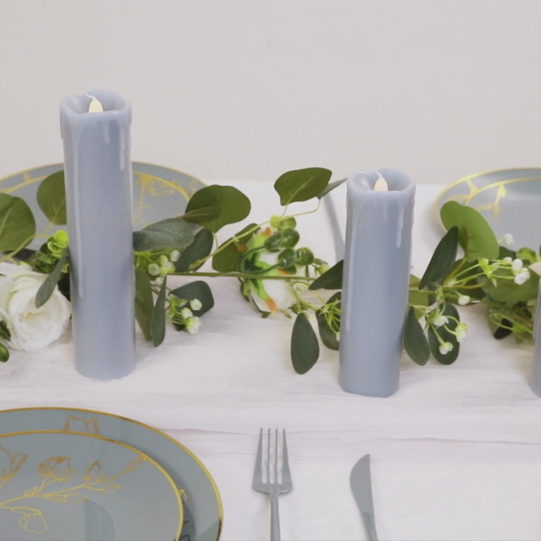 Dusty blue LED drip wax pillar candles on a table with greenery and elegant dinnerware.