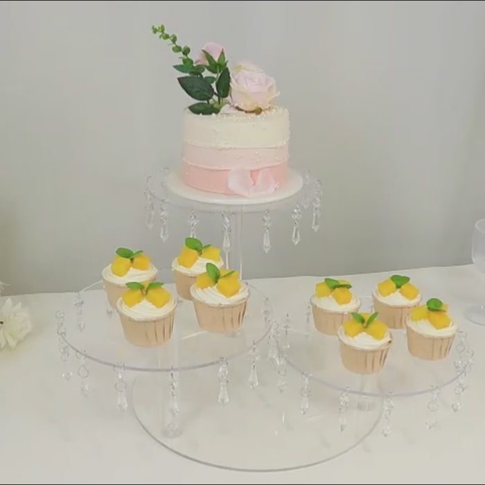Clear acrylic cake stand with hanging crystal pendants displaying cupcakes and a cake.