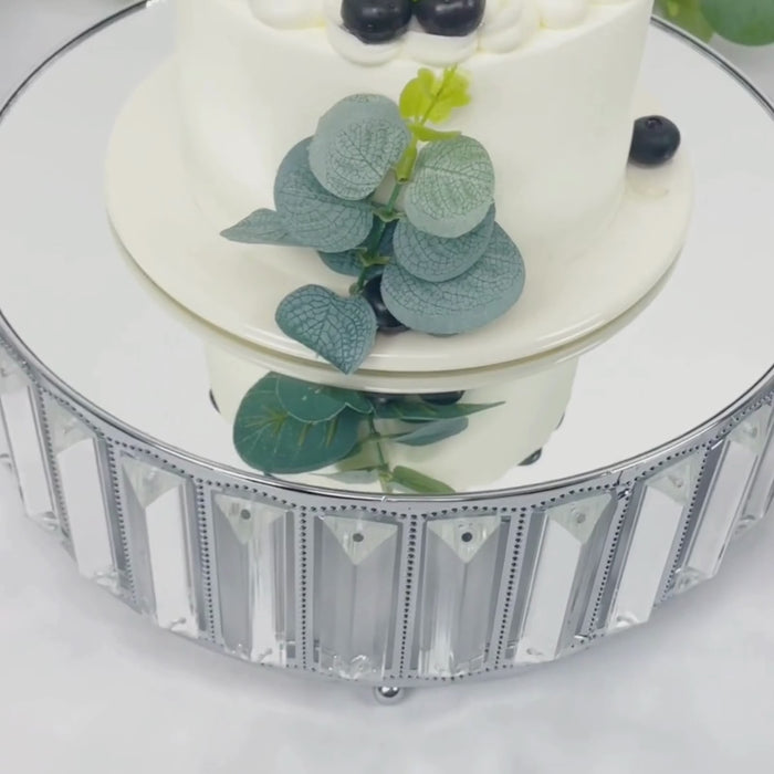 Silver metal crystal cake stand with mirror top, displaying a cake and greenery.