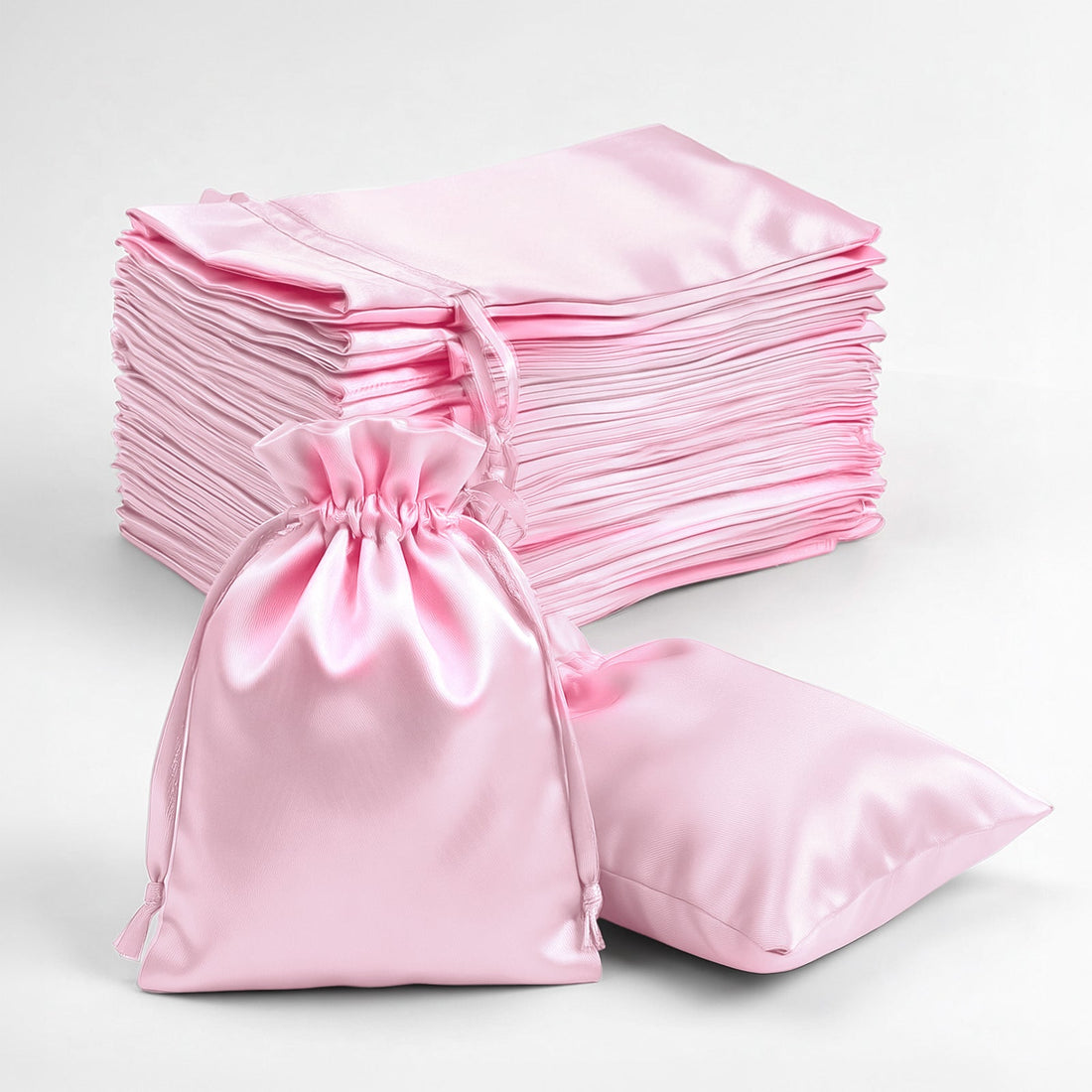 Pink satin drawstring bags stacked with one bag and a pillow on a white background