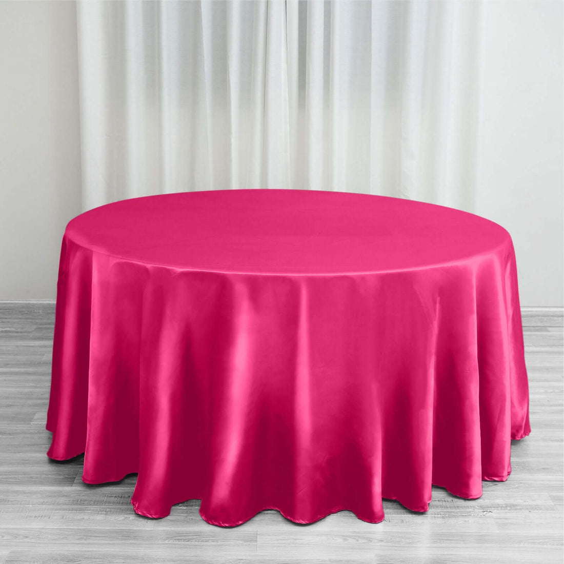 Round pink satin tablecloth on a wooden floor with curtains in the background.