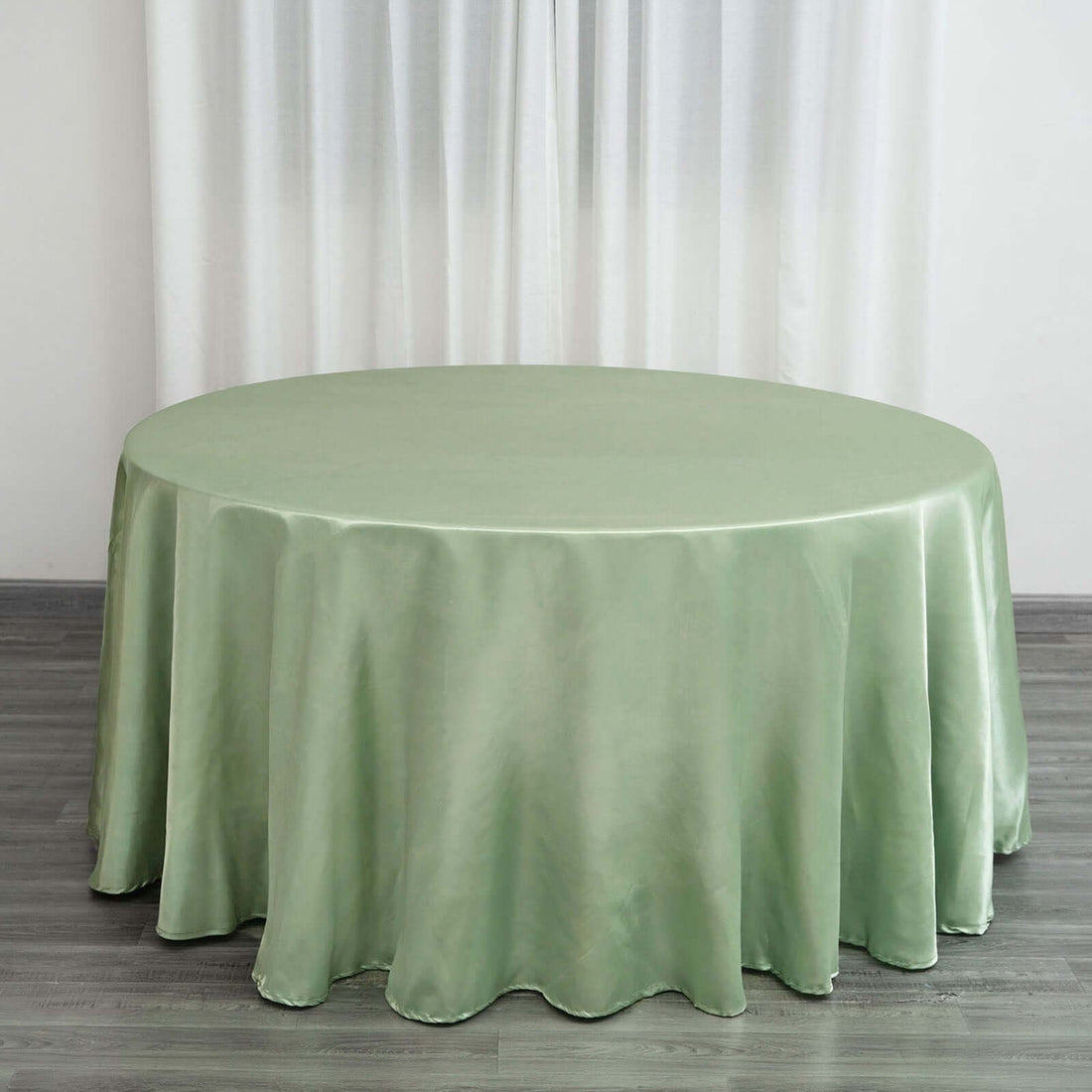 Round green satin tablecloth on a wooden floor with sheer curtains behind it.