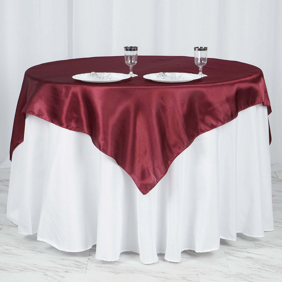 Round table set with burgundy satin tablecloth and white dinnerware on a white backdrop.