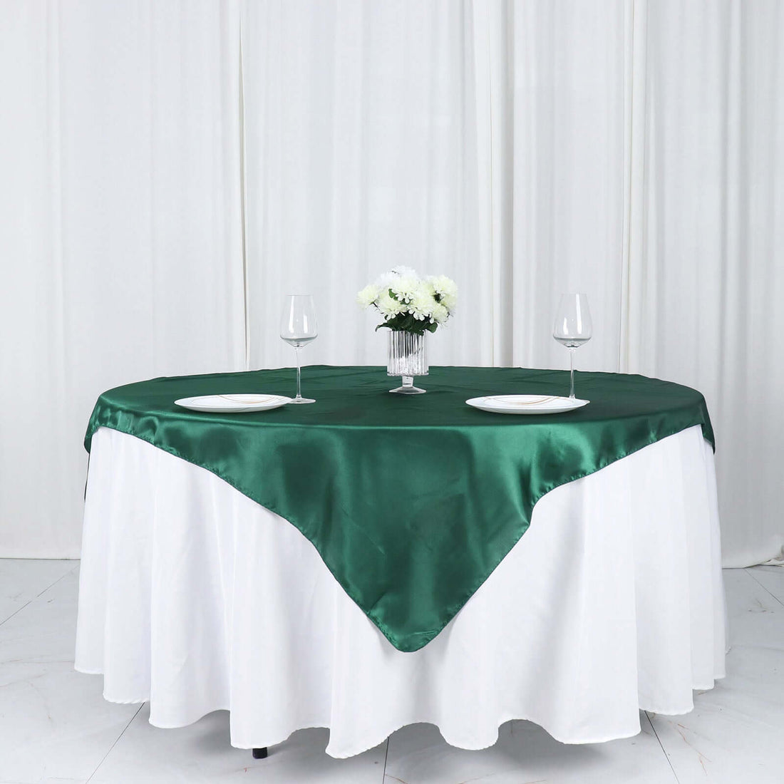 Elegant round green satin tablecloth draped on a white table with wine glasses and flowers.