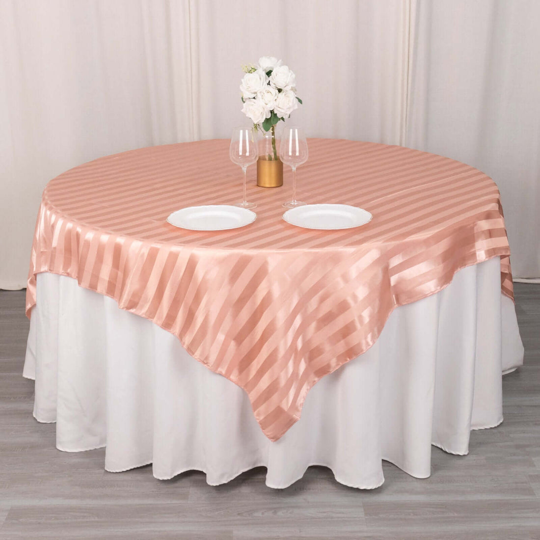 Round peach satin tablecloth draped over a table with white plates and flowers.