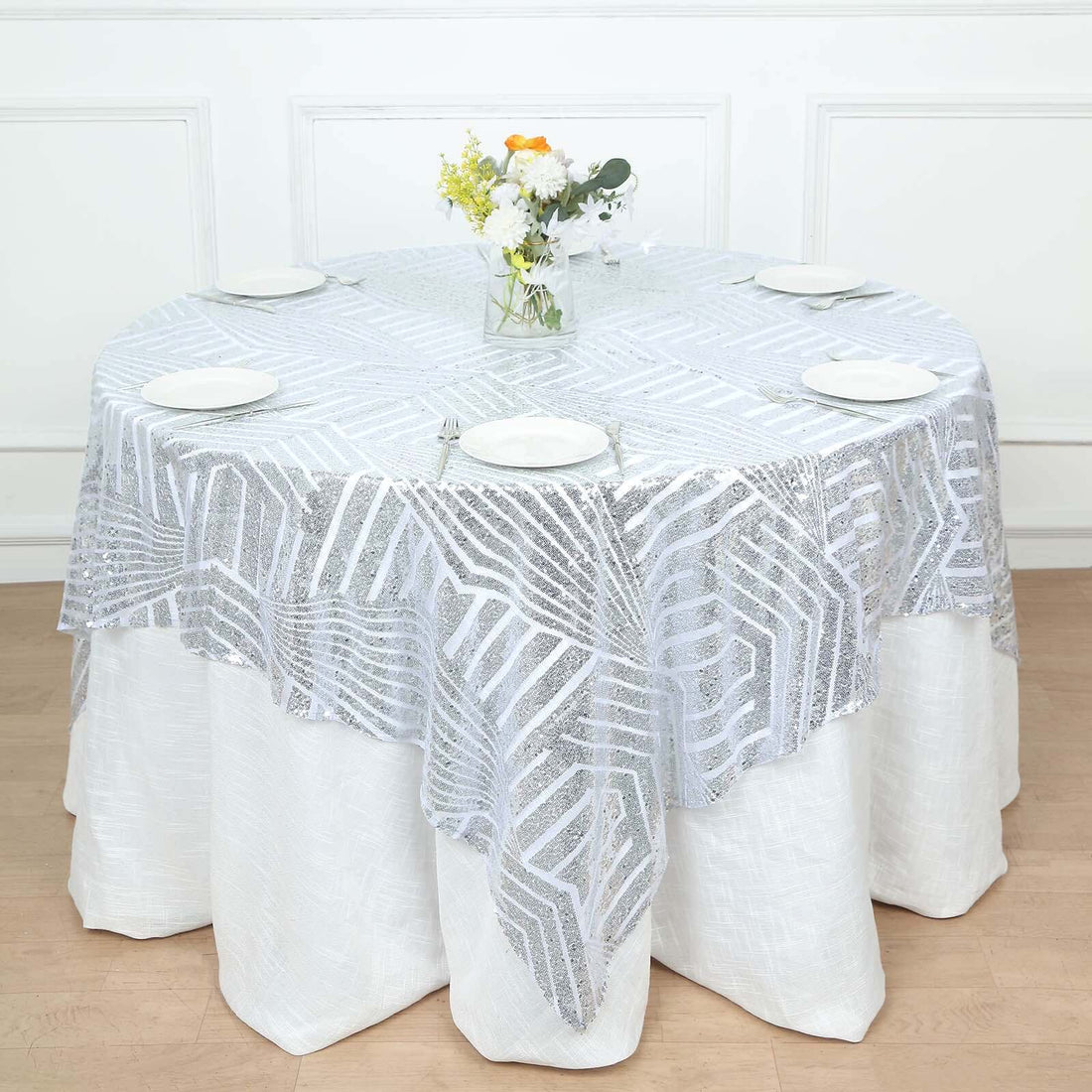 Round table set with a decorative silver lace tablecloth and floral centerpiece.