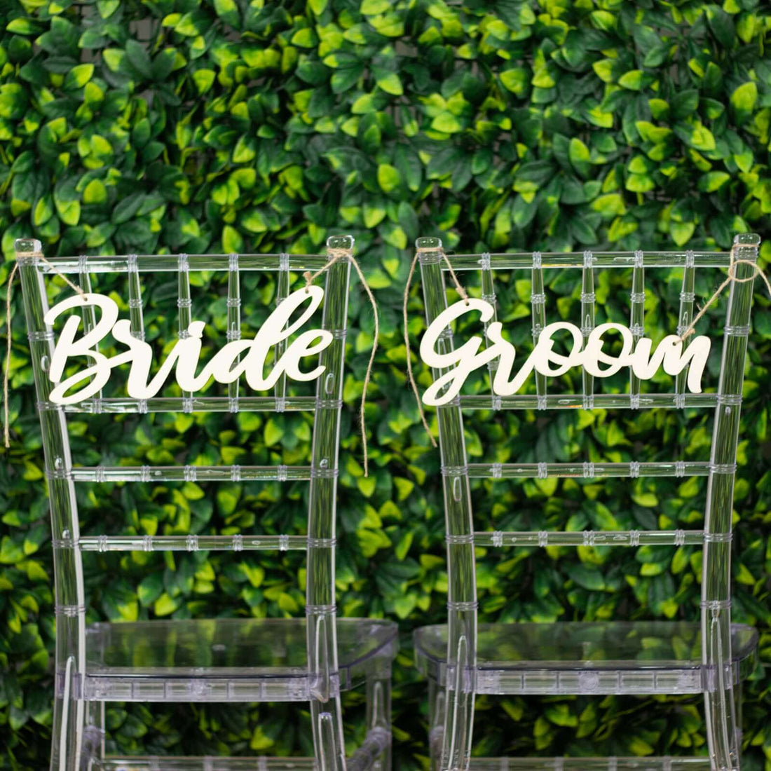 Clear acrylic wedding chairs labeled "Bride" and "Groom" against a greenery backdrop.