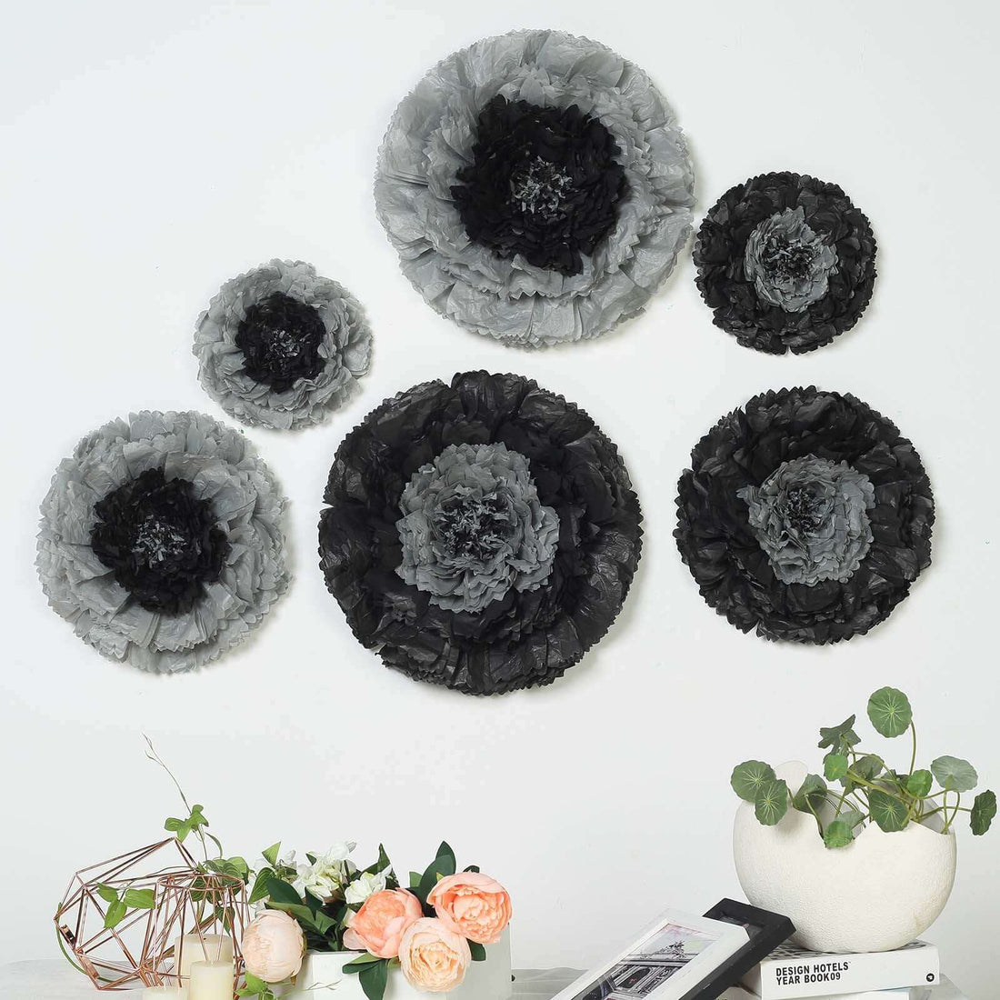 Black and gray decorative paper flowers arranged on a wall with plants in foreground.