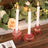 Pink glass candle holders with white candles on a wooden table surrounded by greenery.