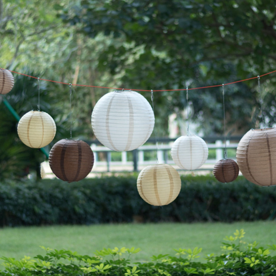 Decorative paper lanterns in various sizes hanging outdoors among greenery.
