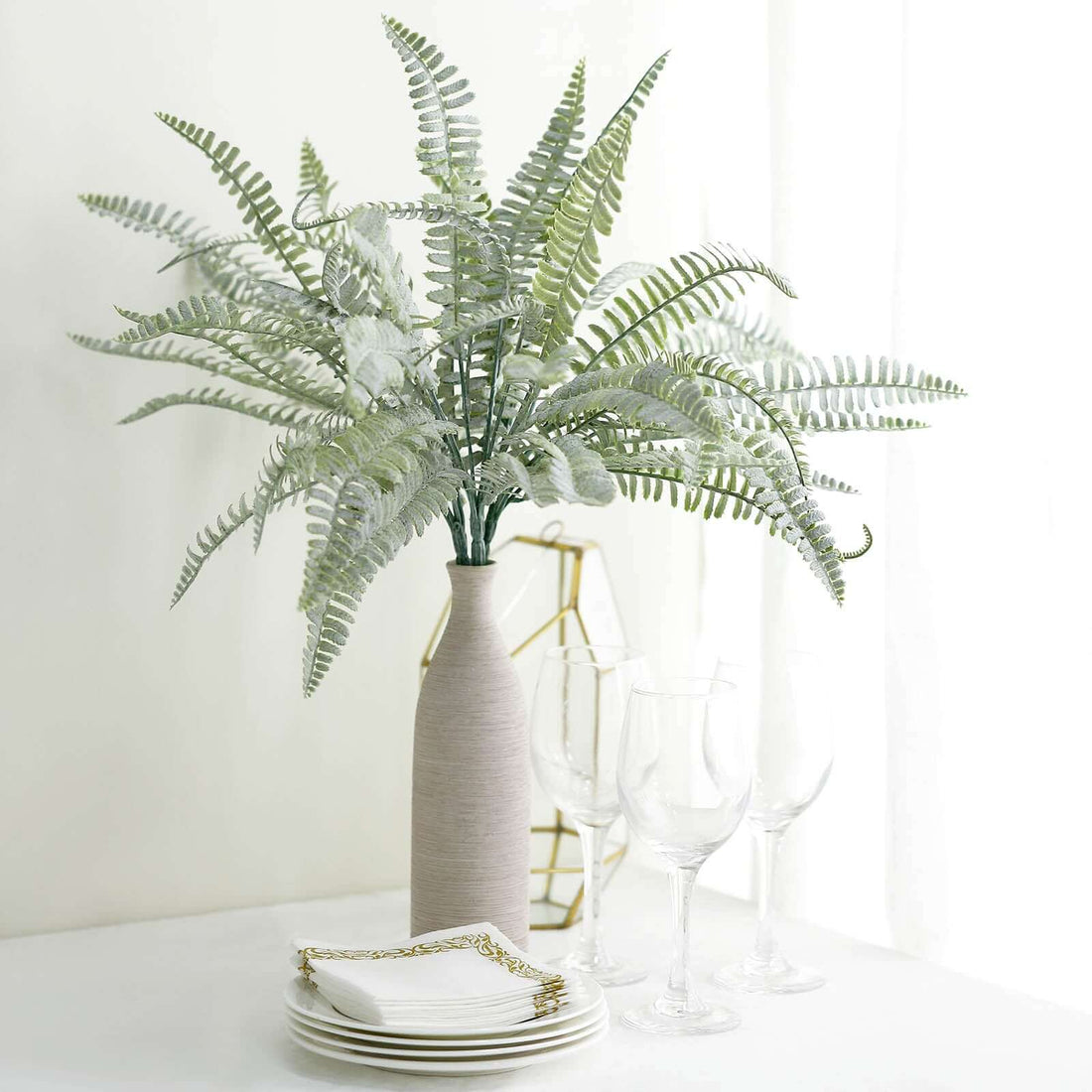 Decorative fern arrangement in a vase with plates and glasses on a table.