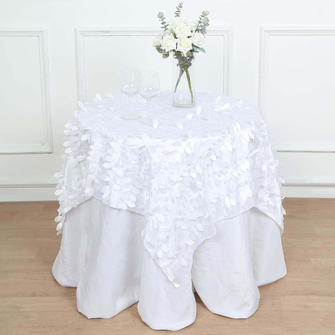 White tablecloth with leaf design draped over a round table with glasses and flowers.