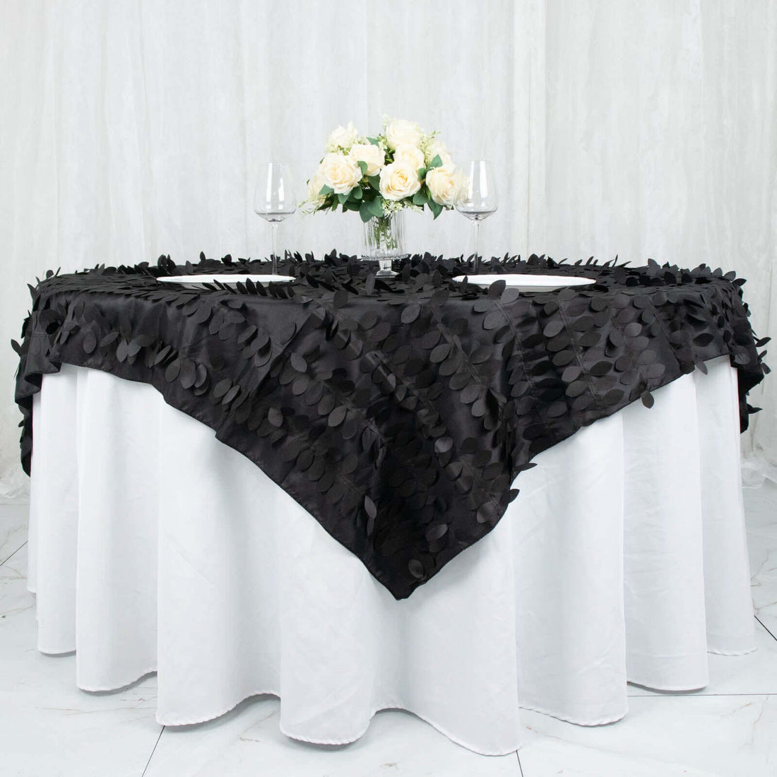 Black leaf-pattern table runner draped over a white table with flowers and glasses.