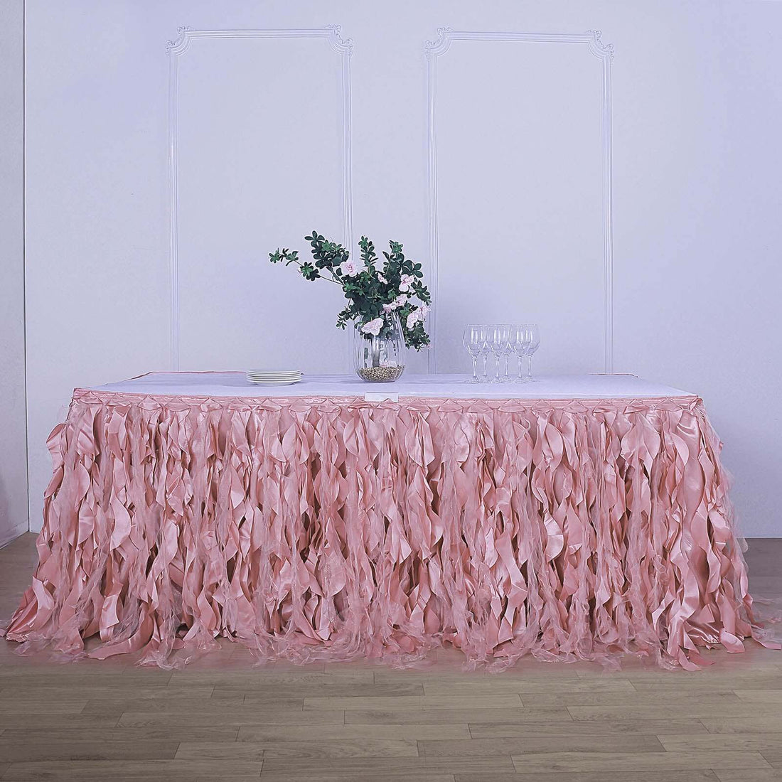 Pink ruffled table skirt with floral centerpiece and glasses on a table.