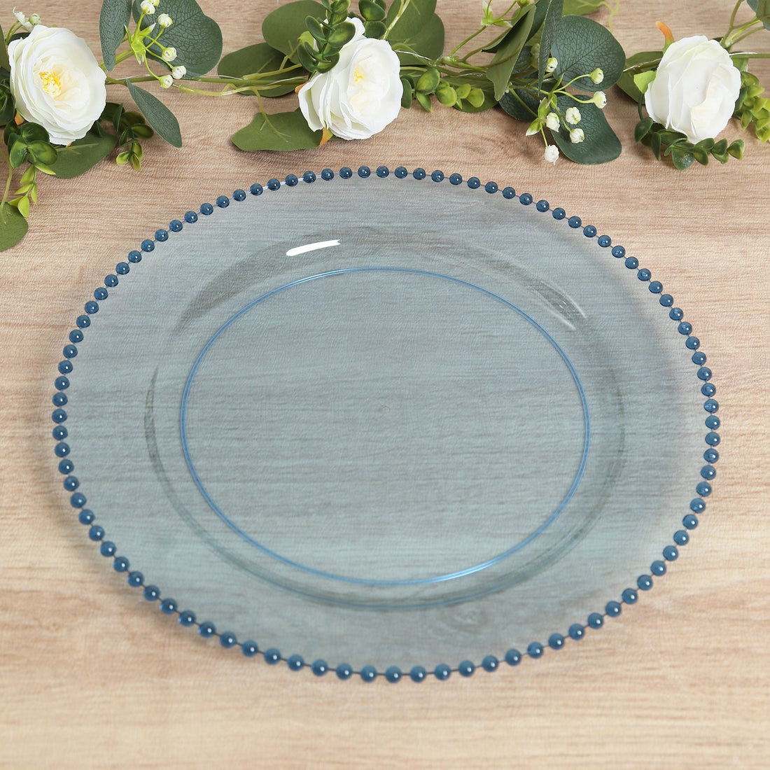 Blue beaded glass charger plate on wooden table with floral accents.