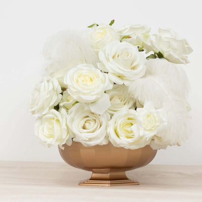 White roses in a gold bowl with feathers on a plain background.