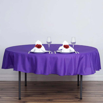 Purple tablecloth on round table set with elegant dinnerware and rose-decorated napkins.