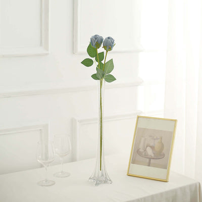 Tall glass vase with blue roses and green leaves on a table.