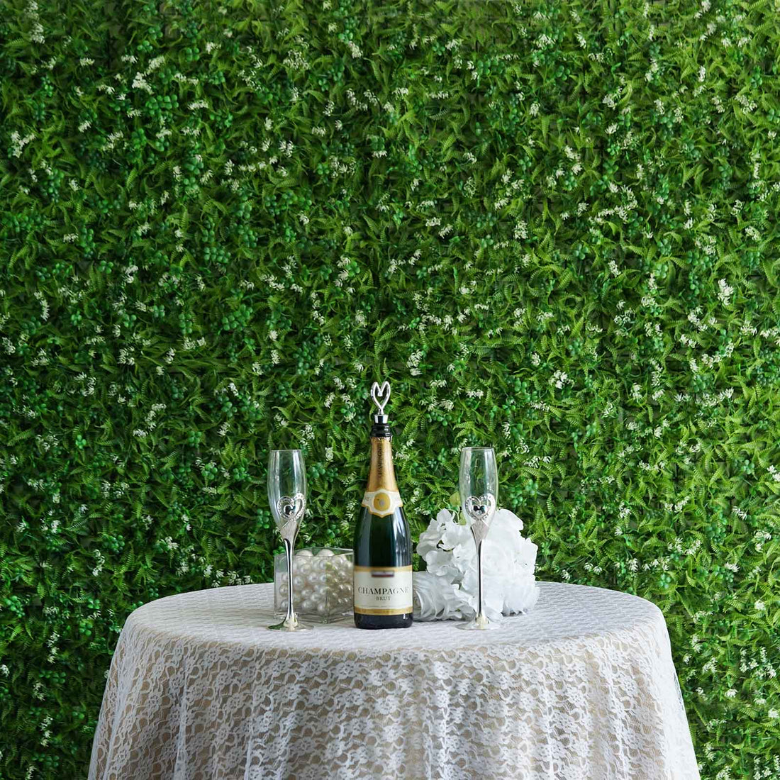 Champagne bottle and glasses on a lace tablecloth with greenery backdrop.