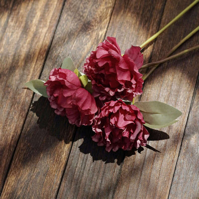 Three deep red artificial peonies with green leaves on a wooden surface.