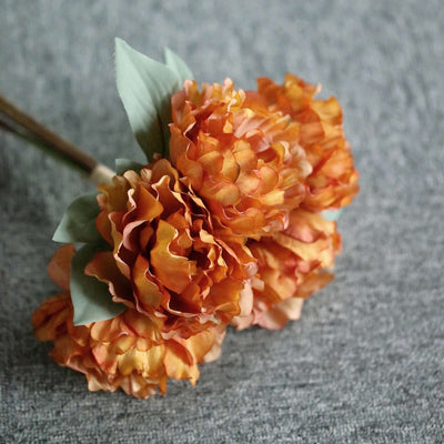 Artificial orange peony bouquet with green leaves on a textured gray background.