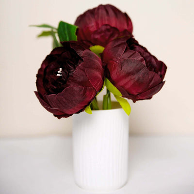 Artificial burgundy peony flowers in a white vase.