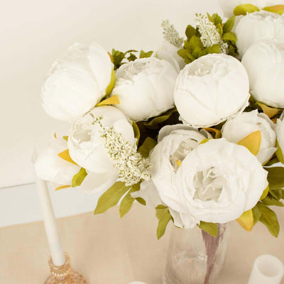 White peony flowers in a glass vase with greenery and candles.