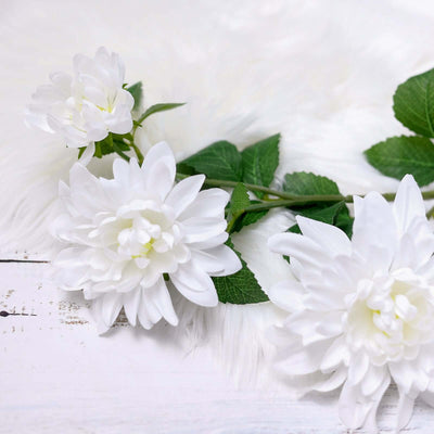 White artificial flowers with green leaves on a soft white surface.