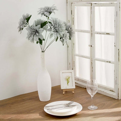 Decorative gray flowers in a white vase on a wooden table with a window backdrop.