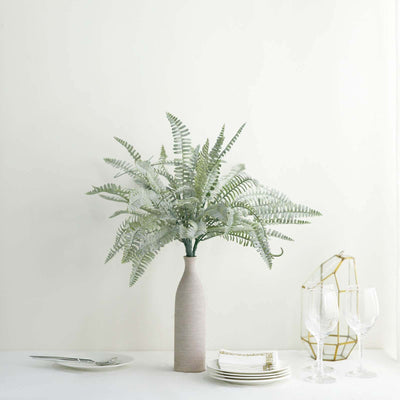 Potted fern arrangement in a minimalist vase on a table with diningware.