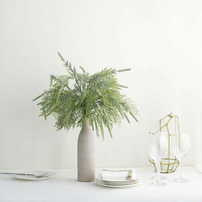 Green foliage arrangement in a vase with plates and glasses on a table.