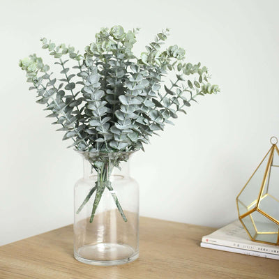 Artificial eucalyptus plant in glass vase on wooden table.
