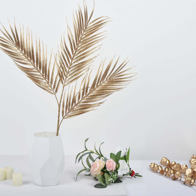 Gold palm leaves in a white vase with pink flowers and candles on a table.
