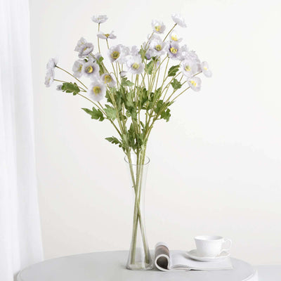 White flowers in a tall glass vase on a table with a cup and magazine.