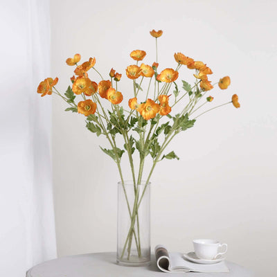 Orange ranunculus flowers in a clear vase on a table with a cup and magazine.