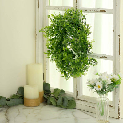 Green artificial wreath hanging by a window with candles and flowers nearby.