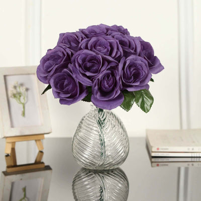 Purple artificial roses in a clear glass vase on a reflective surface.