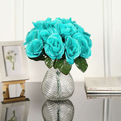 Turquoise faux roses in a glass vase on a reflective surface.