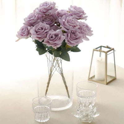 Purple artificial roses in a clear vase with glasses and a lantern.
