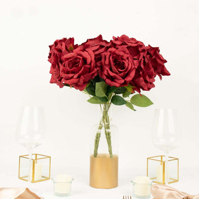 Red artificial roses in a glass vase on a table with wine glasses and candles.