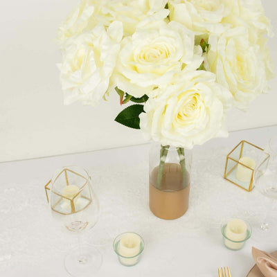 White roses in a glass vase with gold accents, surrounded by candles and glass decor.