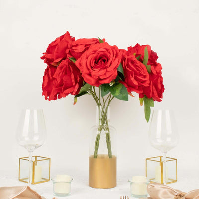 Red artificial roses in a glass vase with gold base, surrounded by elegant table settings.