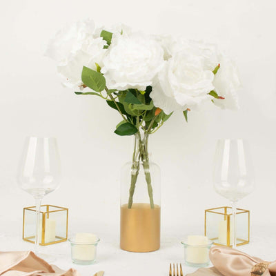 White rose bouquet in glass vase with gold base, surrounded by candle holders and glasses.