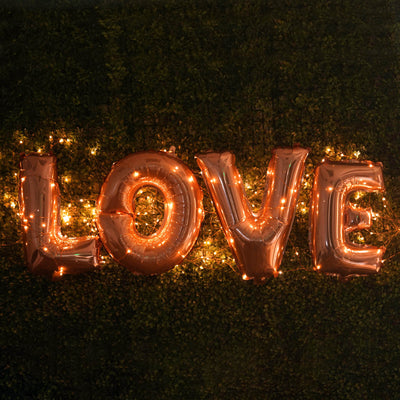 Rose gold "LOVE" balloons with fairy lights on a green backdrop.