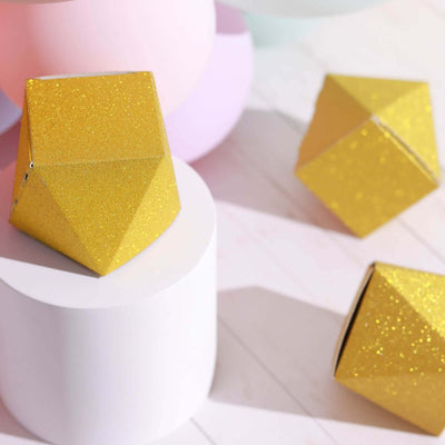 Gold glitter geometric shapes on a white pedestal with pastel balloons in the background.